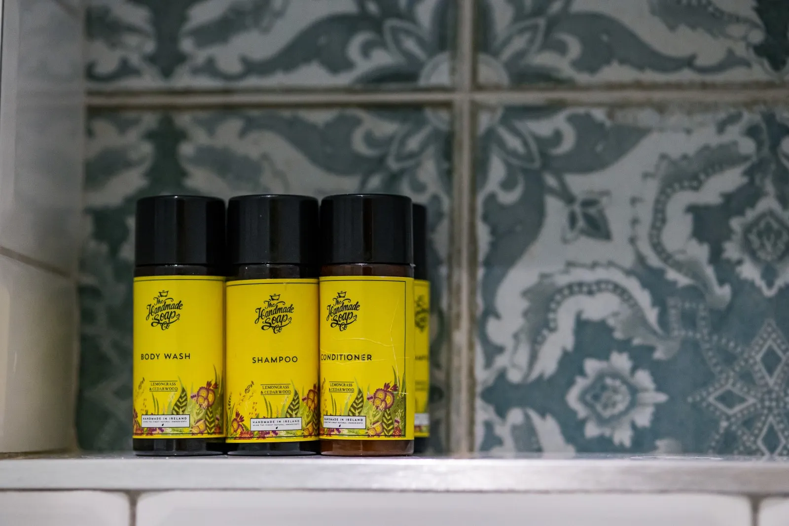 Set of small yellow bottles of body wash, shampoo, and conditioner on a tiled bathroom shelf.
