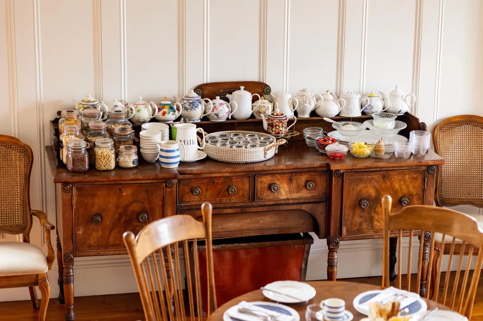 Vintage wooden sideboard with assorted teapots, cereal jars, bowls, and fruit dishes for breakfast.