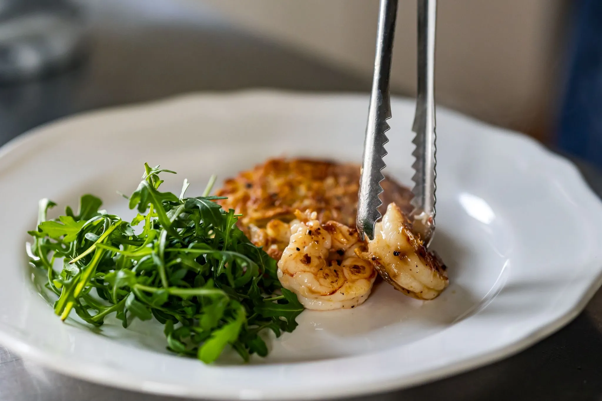 Grilled shrimp being placed on a white plate with fresh arugula and a golden-brown potato pancake.