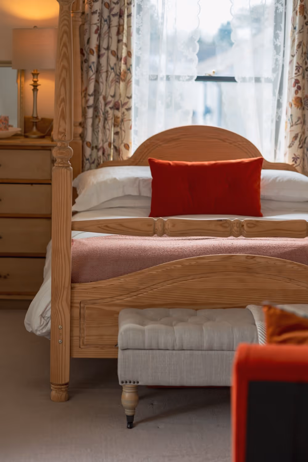 Wooden bed with white linens, red pillow, and floral curtains with sheer white lace curtains in background.