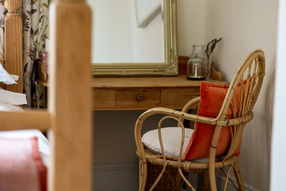 Rattan chair with orange cushions next to a wooden desk with a mirror and glass decor in a cozy corner.