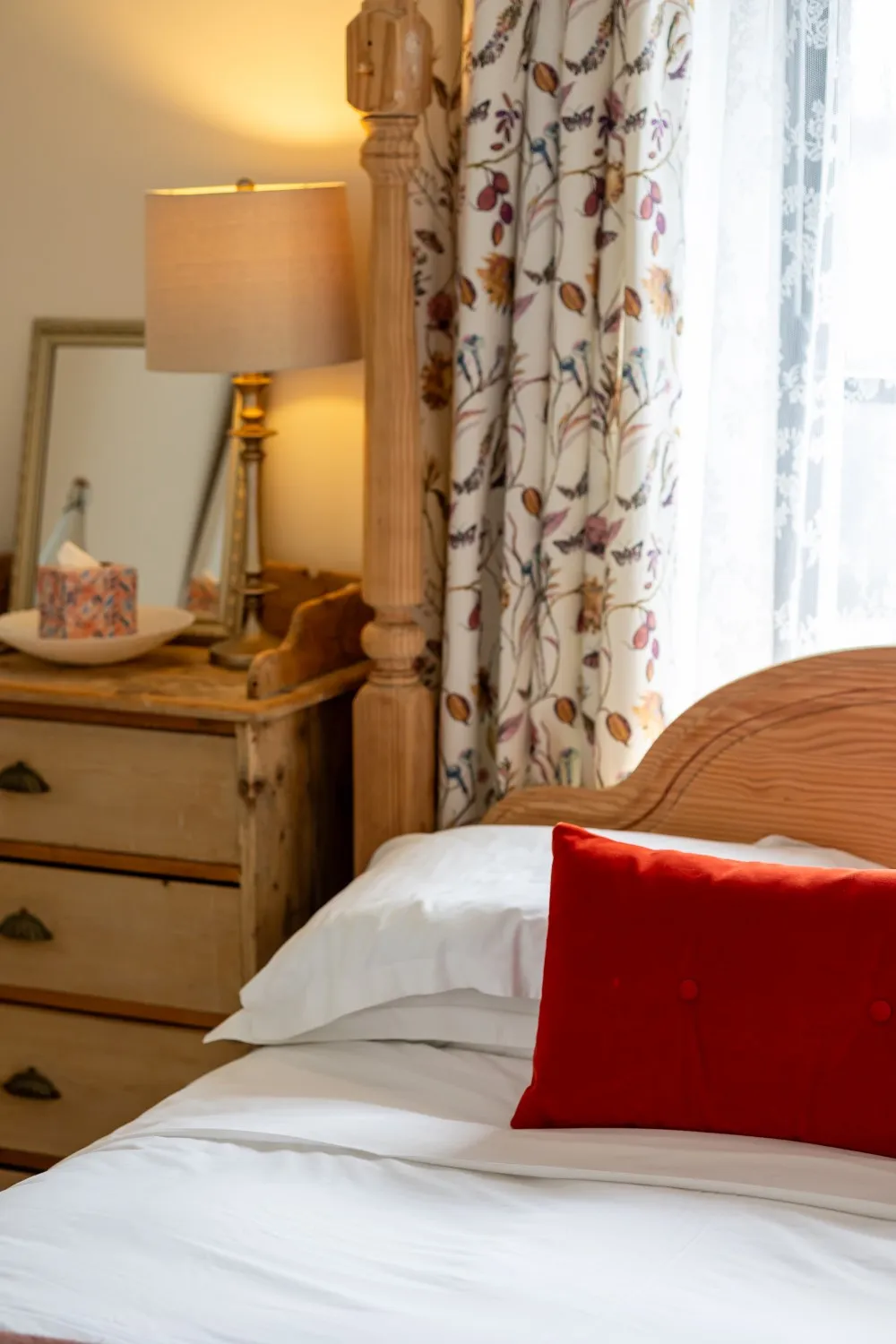 Cozy bedroom corner with wooden bed, white sheets, red pillow, floral curtains, and a lit bedside lamp.
