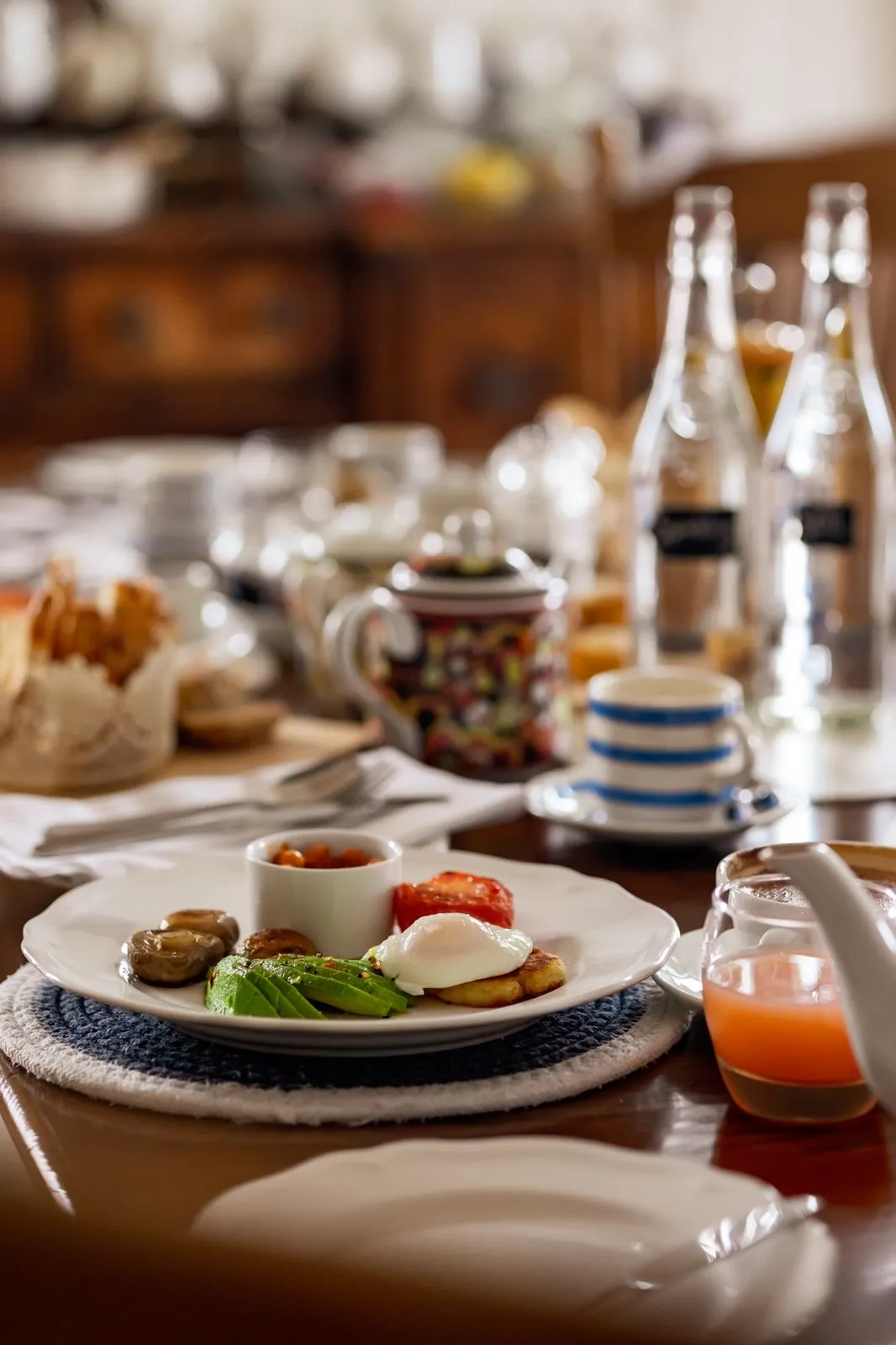 Plate with poached egg, avocado slices, grilled tomato, mushrooms, and baked beans on a dining table.