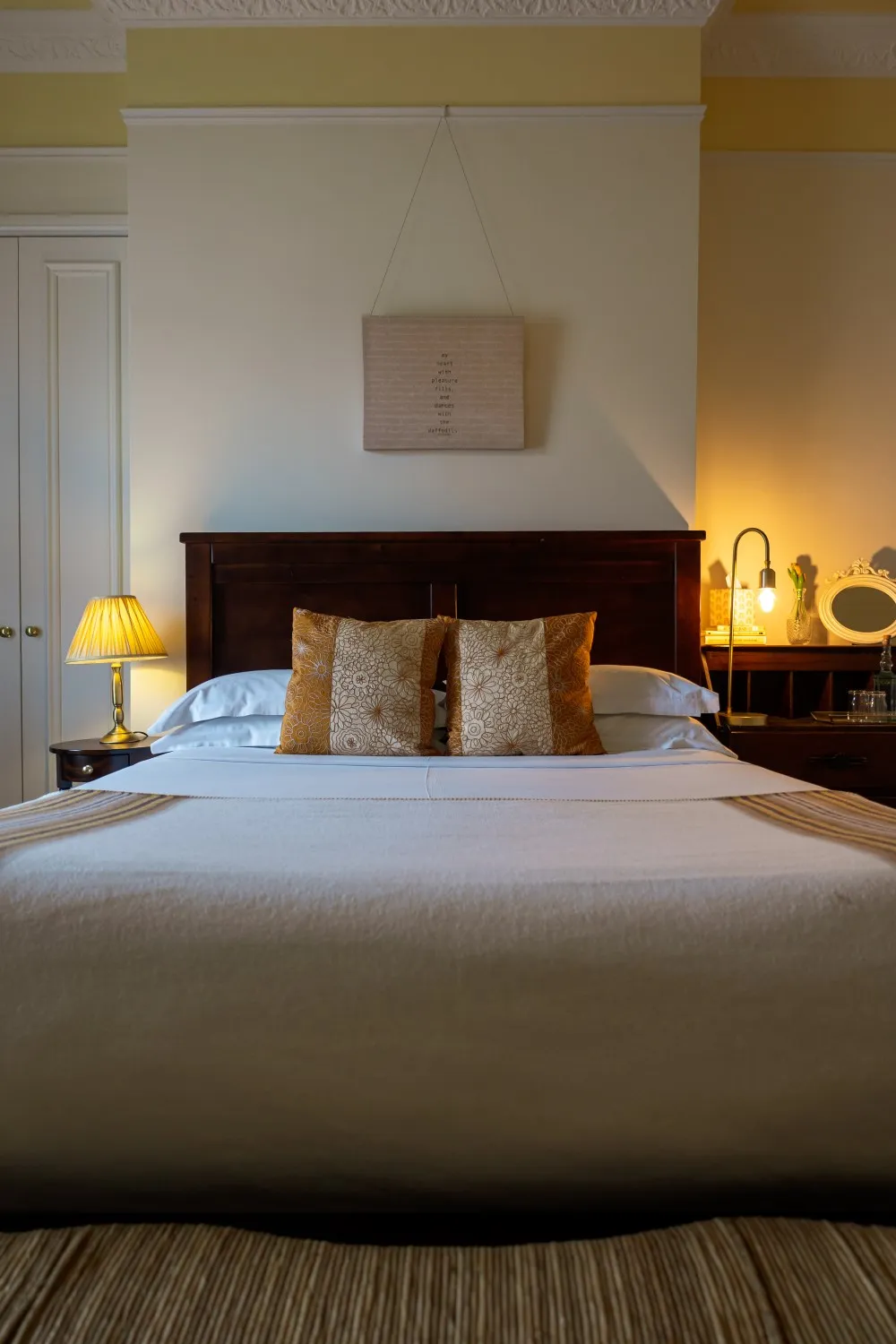 Neatly made bed with white linens, decorative floral pillows, dark wooden headboard, and warm bedside lamps.