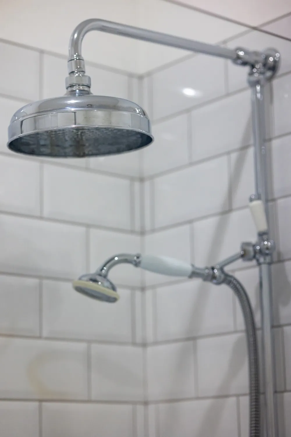 Chrome rain showerhead and handheld shower head mounted on white tiled wall.