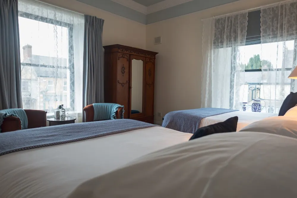 Cozy bedroom with two beds, a wooden wardrobe, two armchairs, and large windows with sheer curtains.