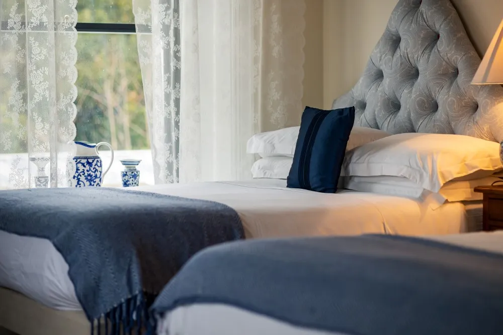 Cozy bedroom with white sheets, blue throws, pillows, and a tufted gray headboard near lace curtains and window.