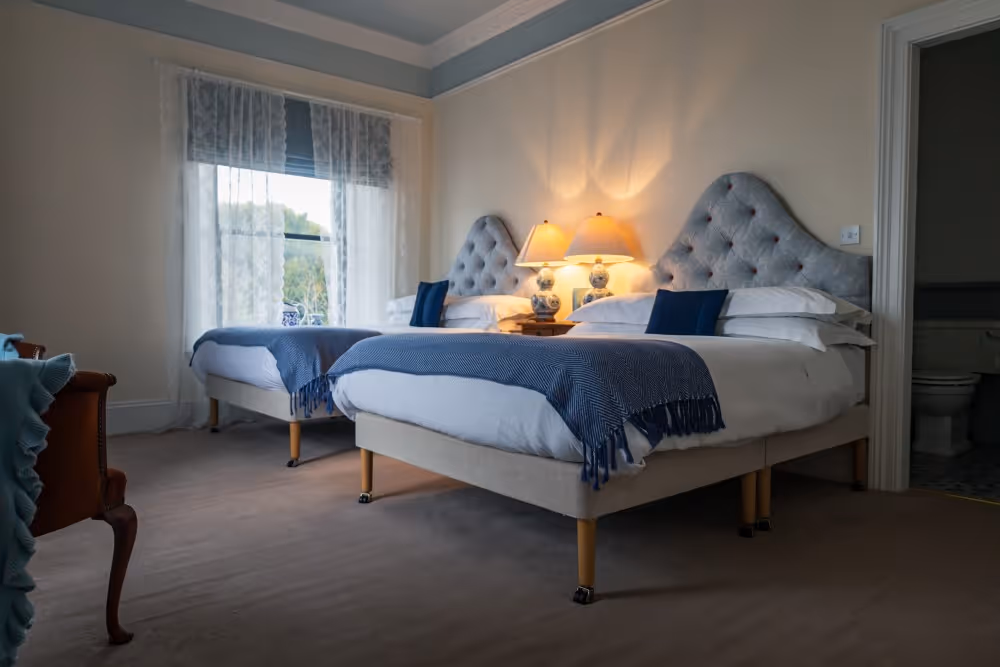 Bedroom with two beds featuring tufted blue headboards, blue throws, and bedside lamps on a shared table.