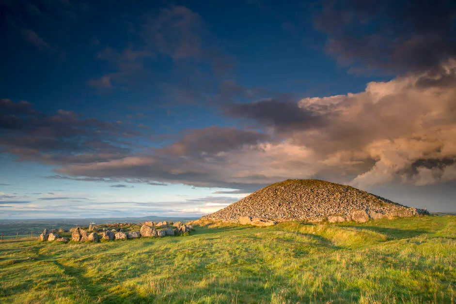 Stone cairn mound on green grass under a moody sky with dark clouds at sunset.