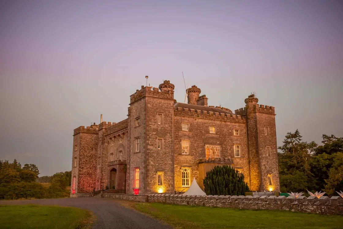 Historic stone castle illuminated at dusk with a curved driveway and lawn in the foreground.