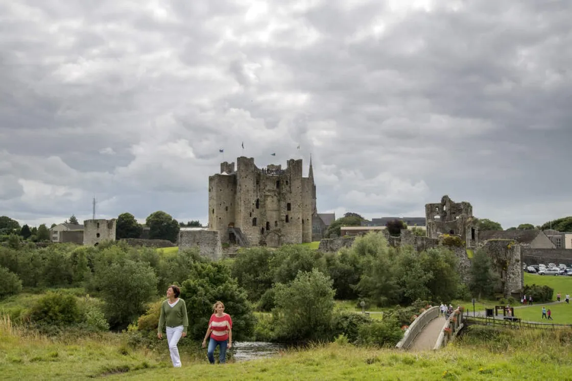Two people walking on a grassy path near a river with a large medieval stone castle in the background under cloudy sky.