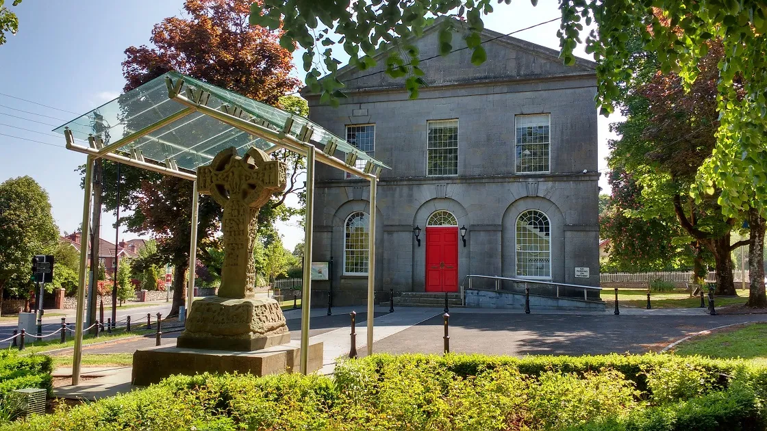 Historic stone high cross under glass shelter with grey courthouse building featuring a red door in the background.