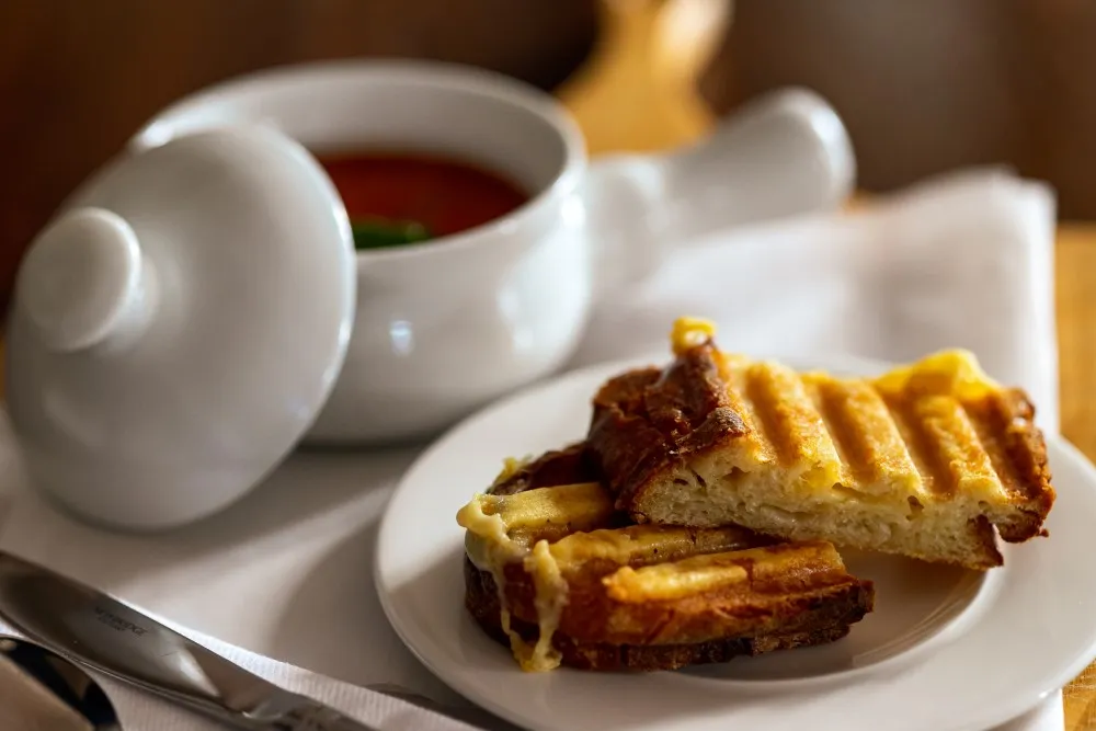 Grilled cheese sandwich halves on a white plate with tomato soup in a white bowl with lid on a table.