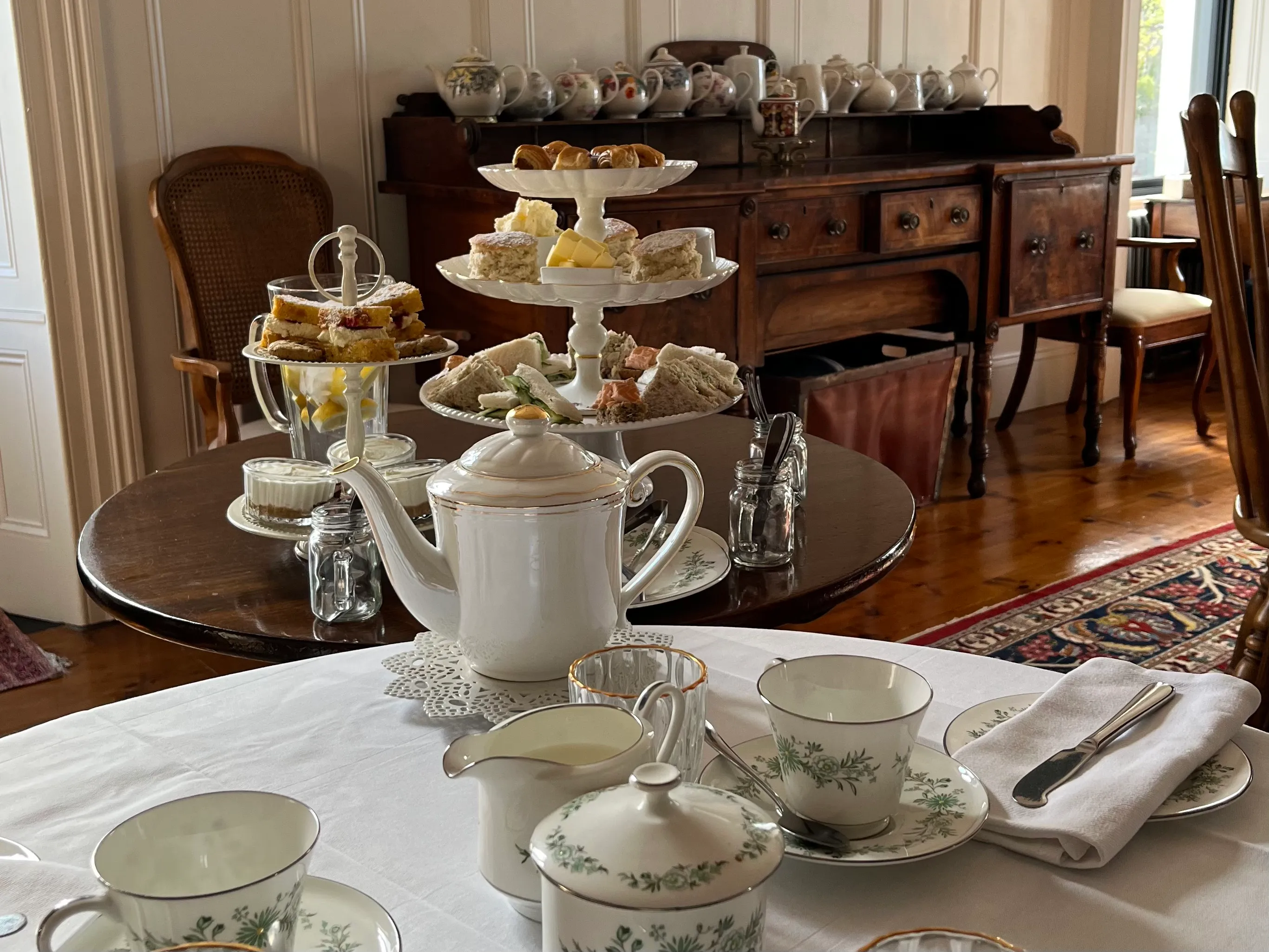 Table set for afternoon tea with teapot, cups, milk jug, sugar bowl, and tiered tray of sandwiches and pastries.