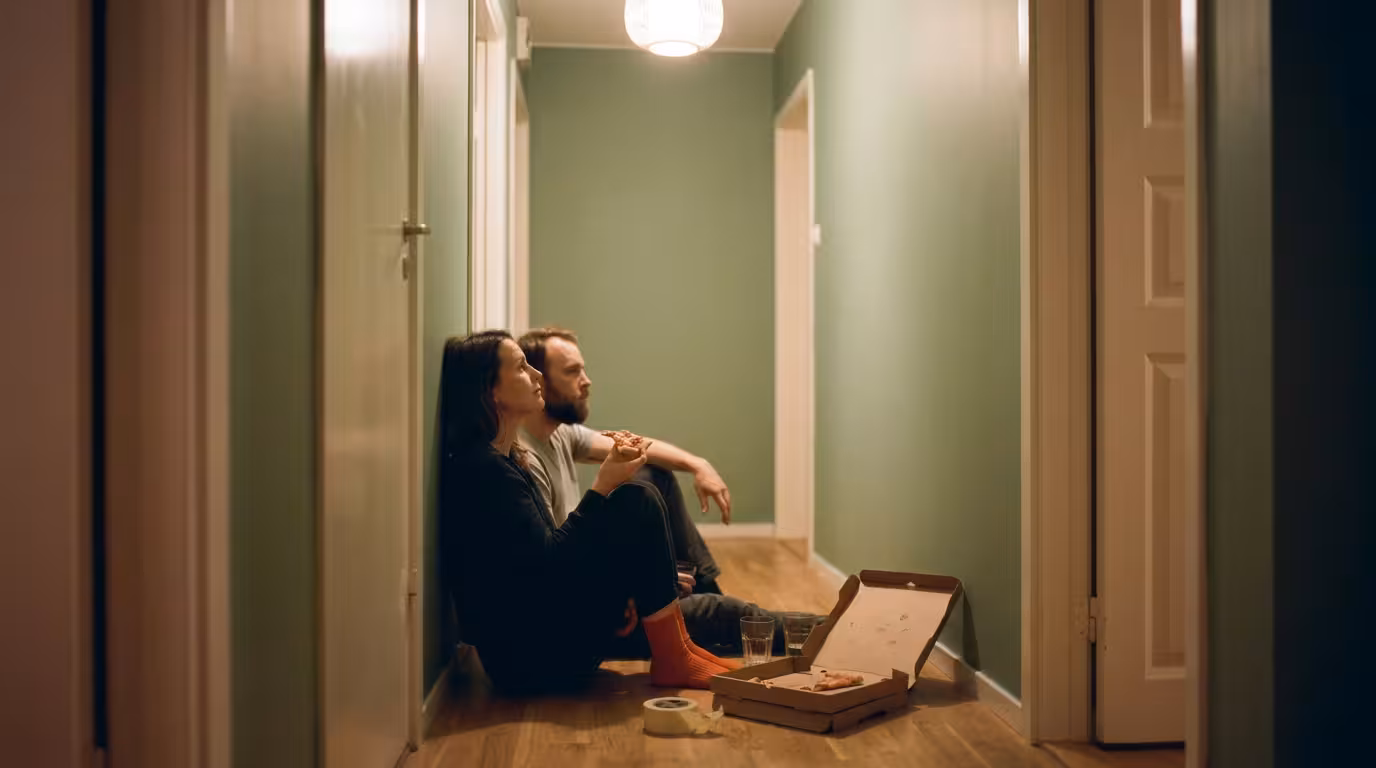 Par sitter på ett hallgolv och äter pizza framför en nymålad vägg