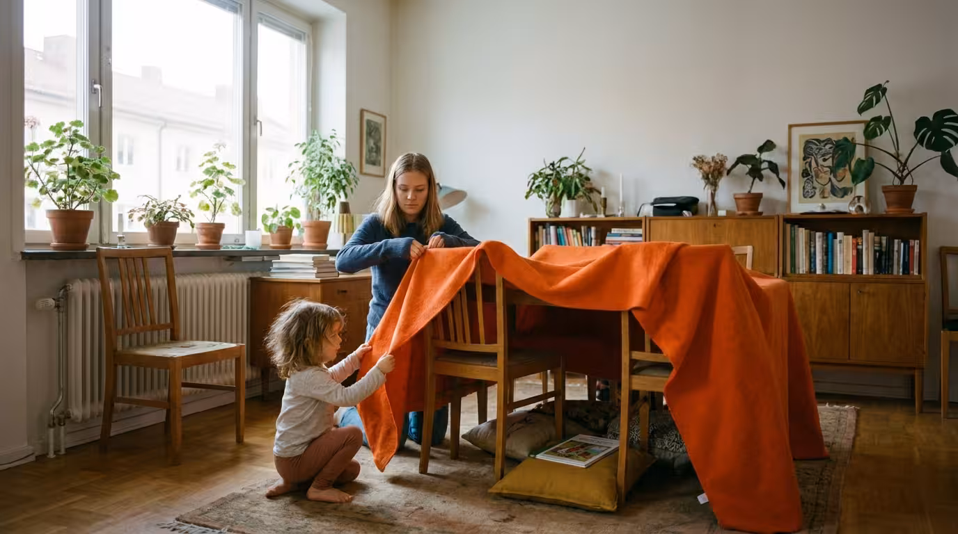 Sjuttonårig tjej och treårig flicka bygger en koja av stolar och en orange filt