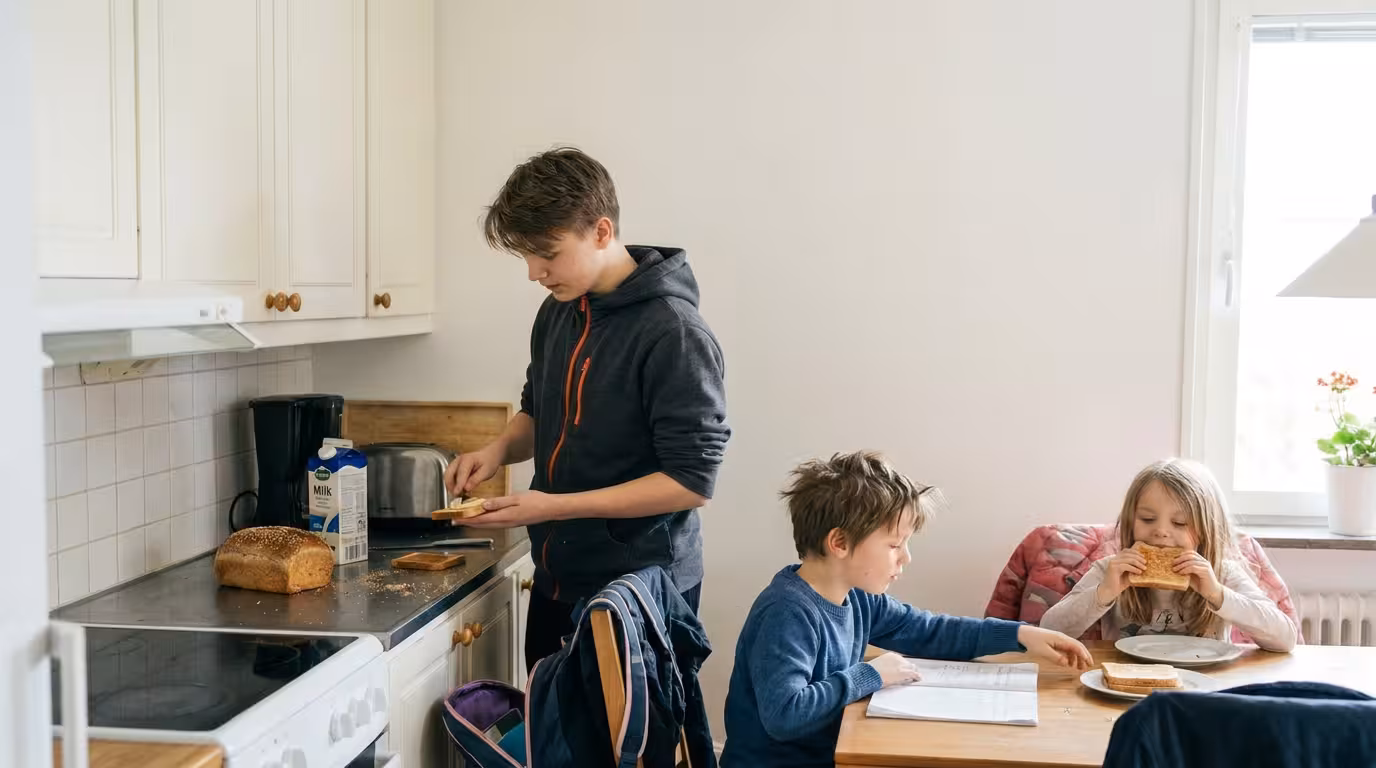 Sjuttonårig kille brer mackor i ett kök medan två skolbarn äter vid bordet
