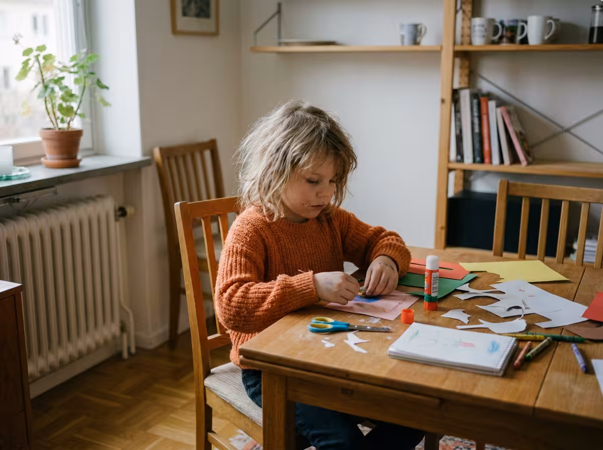 Ett barn sitter vid köksbordet och pysslar med papper, sax och lim