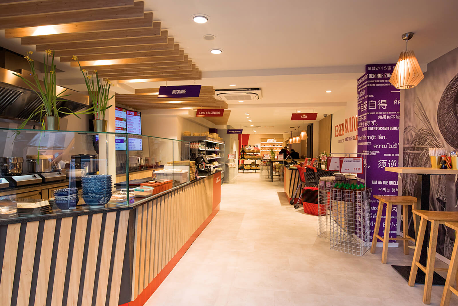 Innenansicht eines modernen, hell beleuchteten Restaurants mit einem langen Holztresen links, blauen Schalen, Glasabtrennung, Pflanzen, digitalen Menüs und mehreren offenen Bereichen für Bestellung, Ausgabe und Kasse, mit Sitzgelegenheiten rechts und dekorativen Elementen an Wänden und Decke.