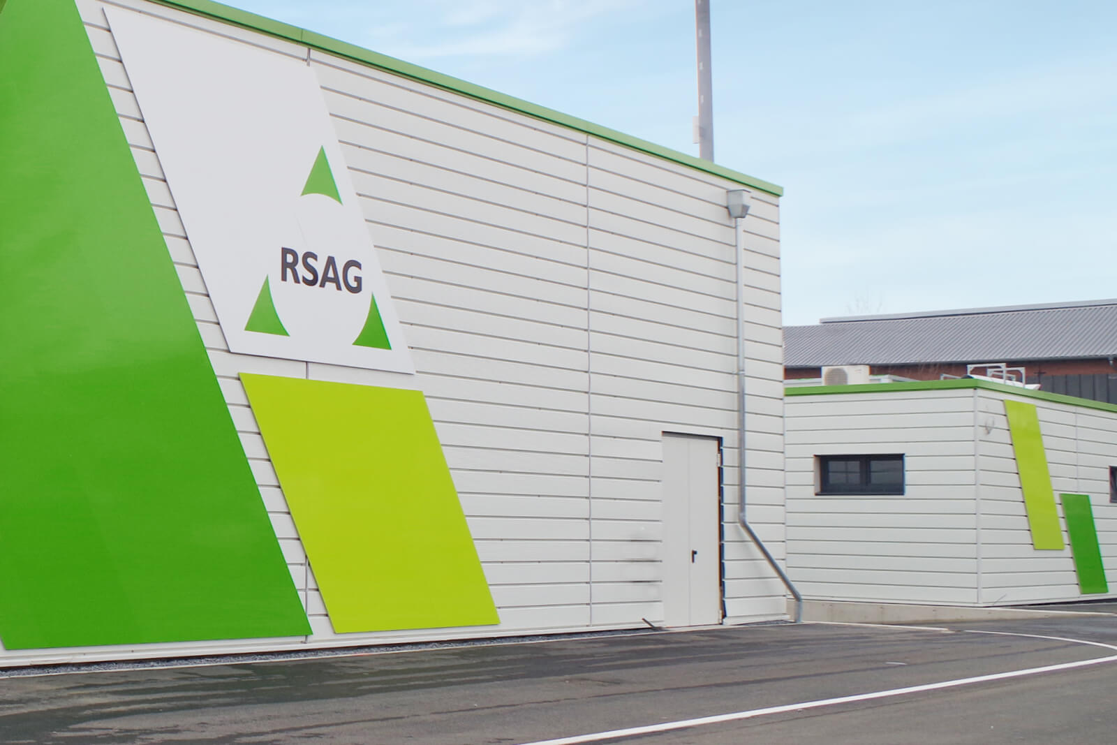 Gebäude mit weißer Fassade und grünen Akzenten, sichtbar das RSAG-Logo auf der linken Seite, mit Türen und Fenstern, davor asphaltierter Bereich.