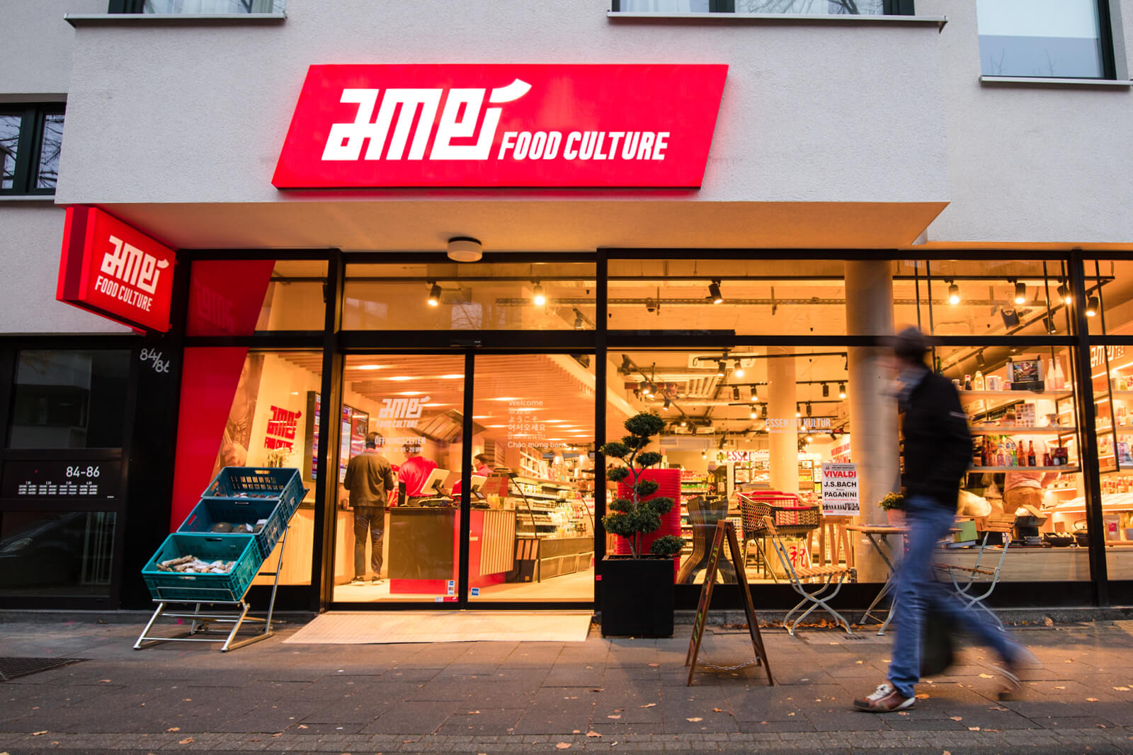 Außenansicht des Lebensmittelgeschäfts AME FOOD CULTURE mit roten Leuchtschildern, einem Verkaufsständer mit Körben rechts vom Eingang und einem Kunden, der den Gehweg entlanggeht.