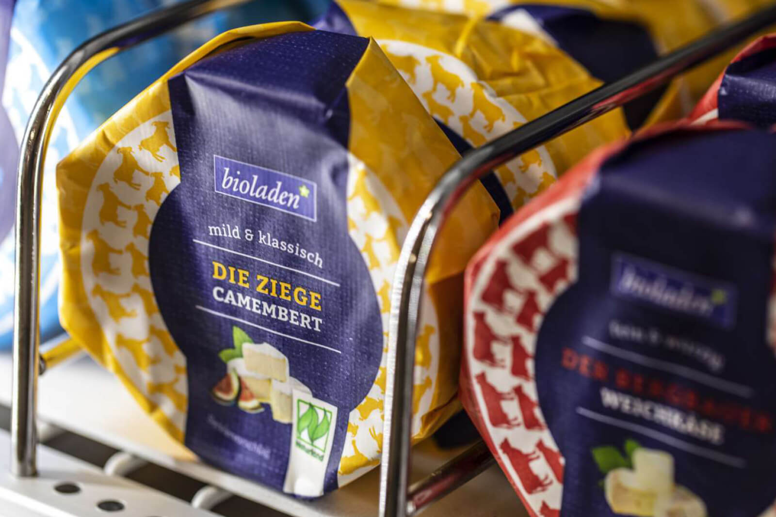 Verpackungen von Bioladen-Camembert-Käse in verschiedenen Sorten, darunter mild & klassisch Die Ziege Camembert, in einem Metallregal im Kühlbereich.