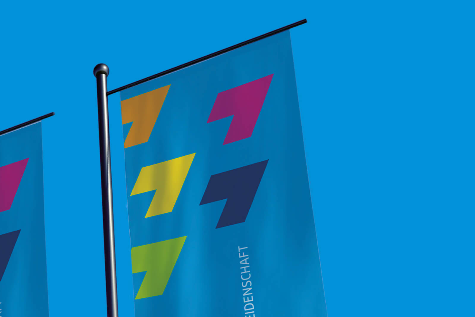 Blaue Fahnen mit farbigen geometrischen Pfeilen in Orange, Pink, Gelb, Dunkelblau und Hellgrün an schwarzen Fahnenstangen vor klarem blauem Himmel.