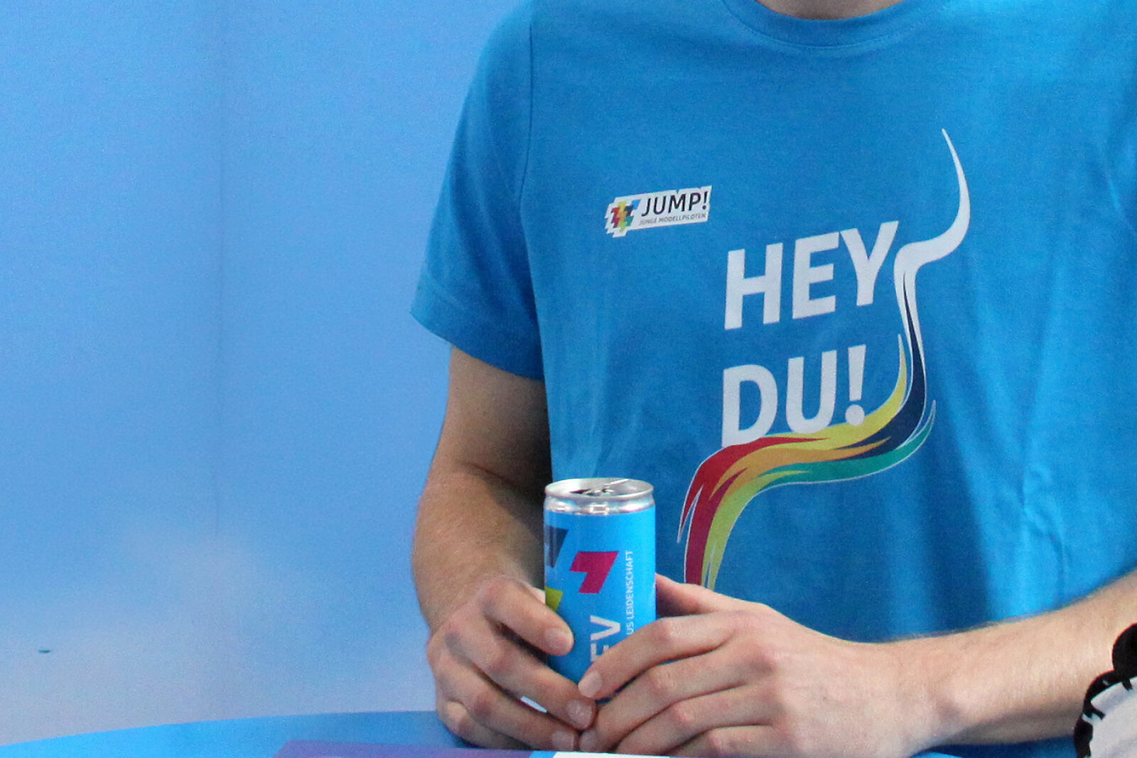 Person trägt blaues T-Shirt mit Aufdruck 'HEY DU!' und mehrfarbigem Design, hält eine blaue Getränkedose mit buntem Logo in beiden Händen auf einem blauen Tisch.