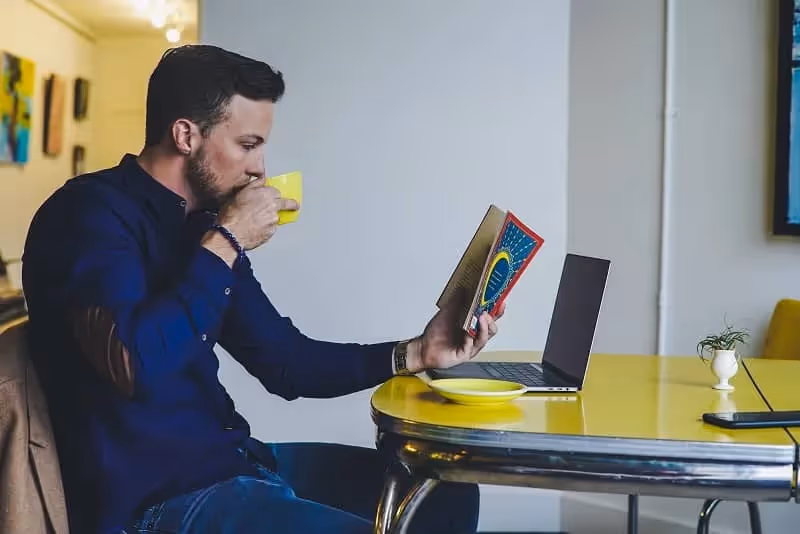 homme qui lit un livre en buvant un café dans une tasse jaune, il est aussi assis devant son ordinateur posé sur une table jaune