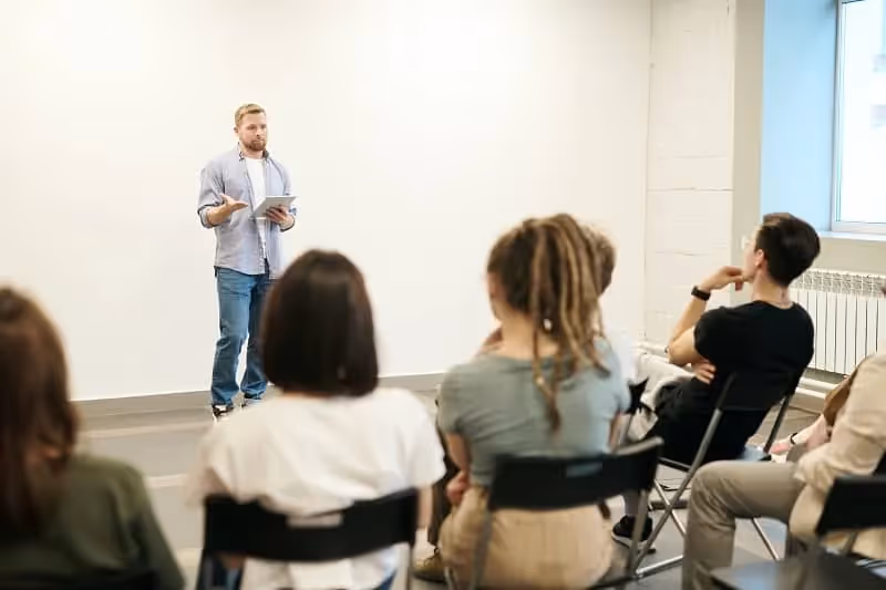 homme qui se présente en anglais dans une salle avec mur blanc et avec autour un autre groupe de personne qui l'écoute attentivement