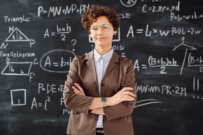 professeure de mathématiques cheveux courts qui se tient devant un tableau rempli de calculs dans une salle de classe