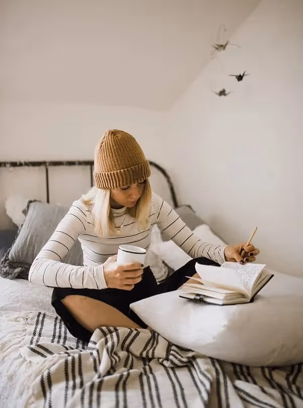 fille avec un bonnet jaune qui étudie assise sur son lit et qui écrit sur un carnet