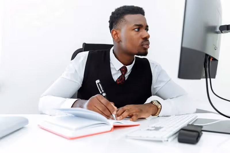 homme installé dans une pièce blanche qui écrit dans un cahier tout en fixant l'écran de son ordinateur