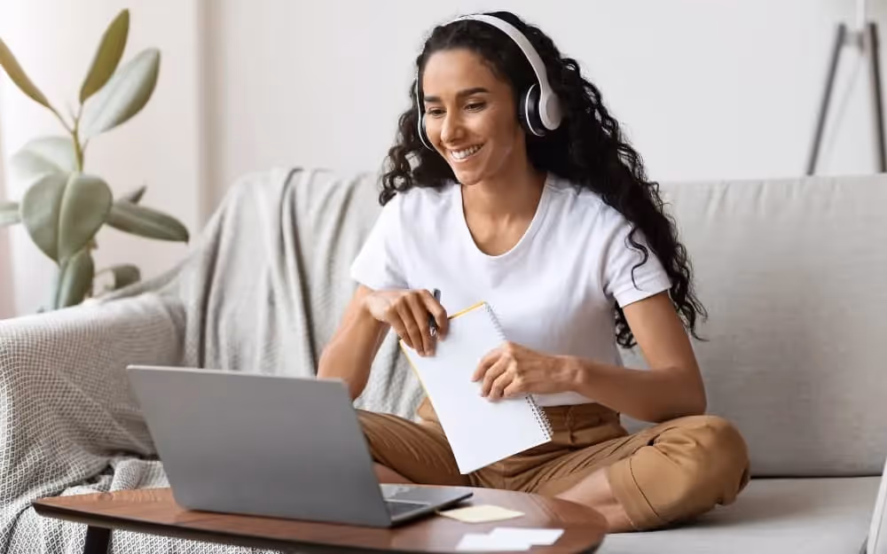 Femme qui suit un cours d'anglais assise dans son canapé avec un casque dans les oreille et un grand sourire pour apprendre l'anglais afin de voyager 