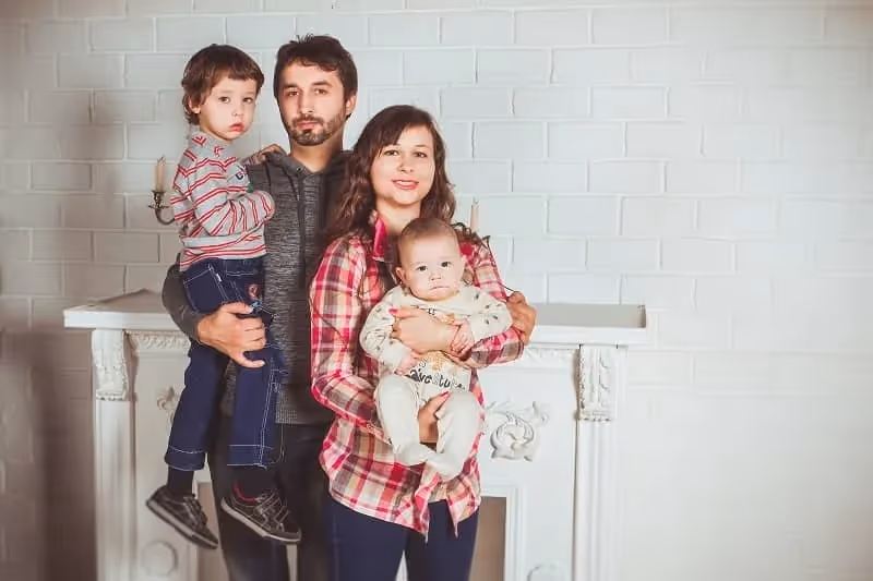 parents avec deux enfants posant pour une photo avec comme fond un mur briqué blanc et une cheminée