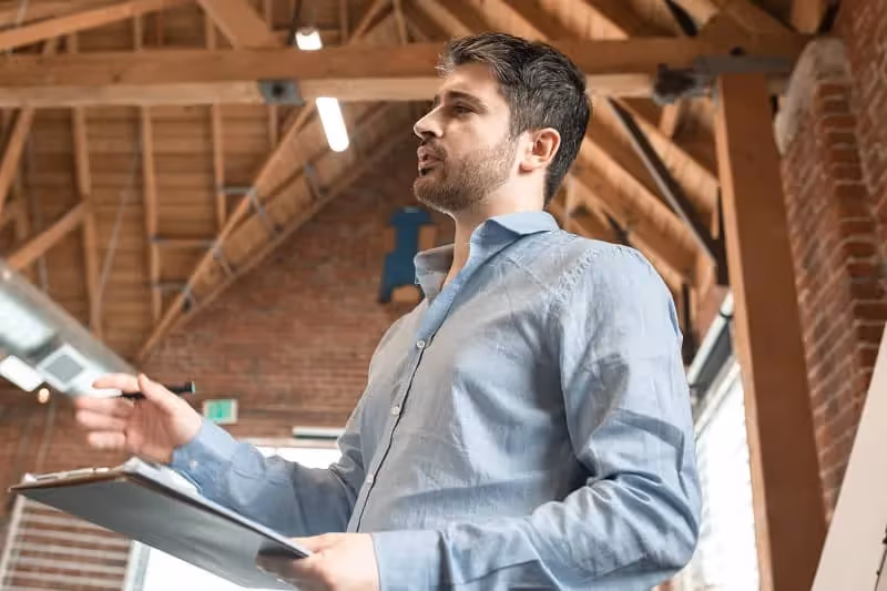 homme barbu habillée d'une chemise bleu ciel explique quelque chose à quelqu'un en tenant un dossier en main