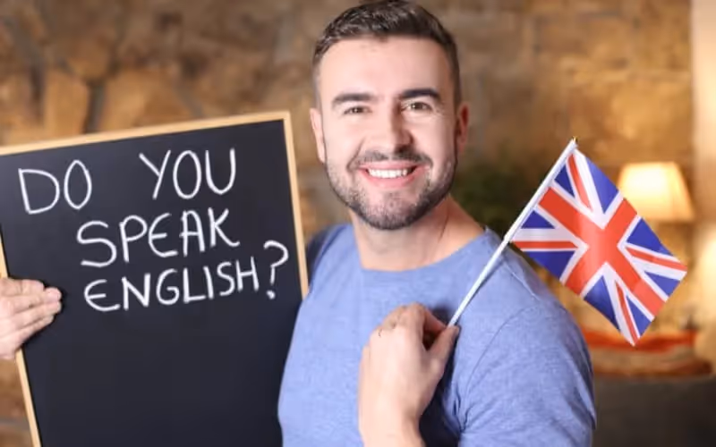 Professeur d'anglais souriant qui tient un tableau sur lequel il est écrit "do you speak englsh", il tient également un drapeau du royaume unis