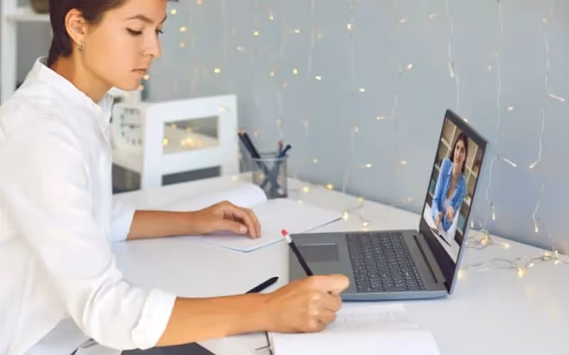 Jeune femme qui suit un cours en ligne sur les adjectifs en anglais dans sa chambre lumineuse sur son bureau blanc avec des guirlandes lumineuses qui descendent le long du mure