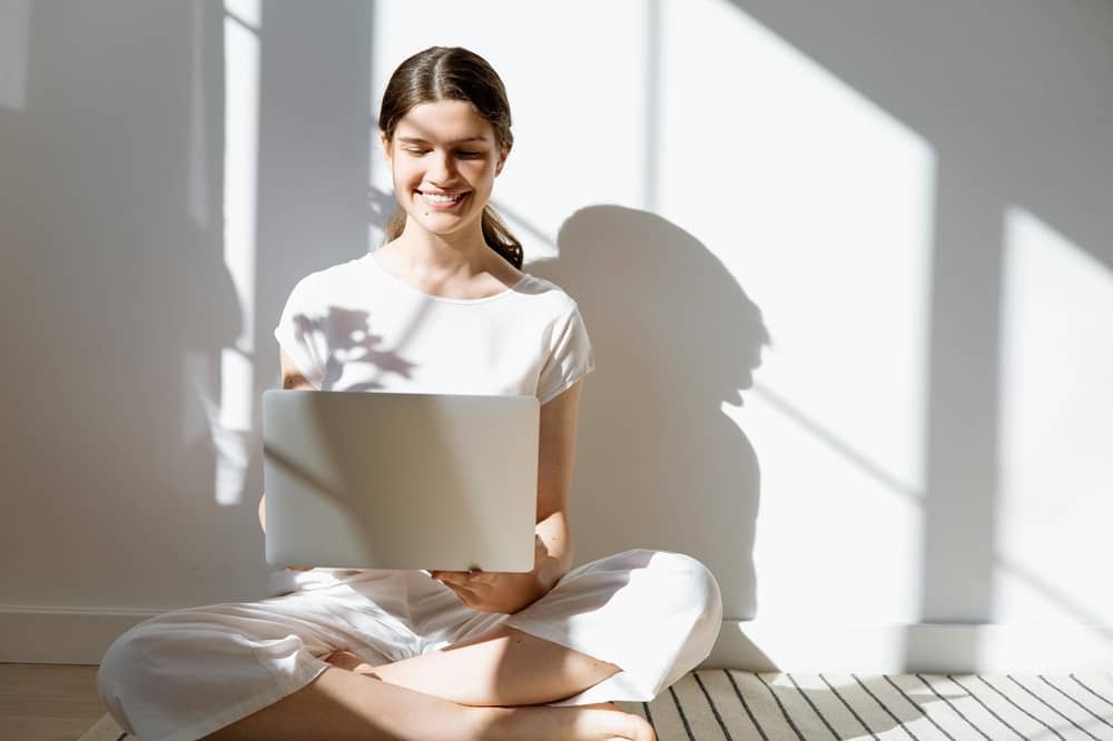 jeune femme qui étudie la liste des 105 verbes à particules en anglais sur son ordinateur assise en tailleur sur le sol