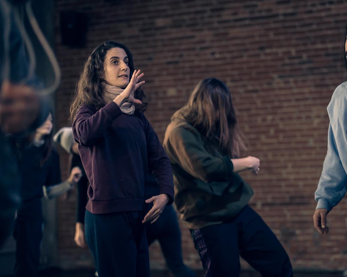 Une photo d'une salle remplie de gens qui dansent. Au centre, Meryem Alaoui est debout et regarde vers l'avant en faisant un mouvement avec ses mains. Elle porte un pull violet foncé, un pantalon bleu foncé et un épais foulard enroulé autour du cou. Derrière elle, une personne portant un pull vert foncé et un pantalon noir se balance, ses cheveux couvrant son visage. 