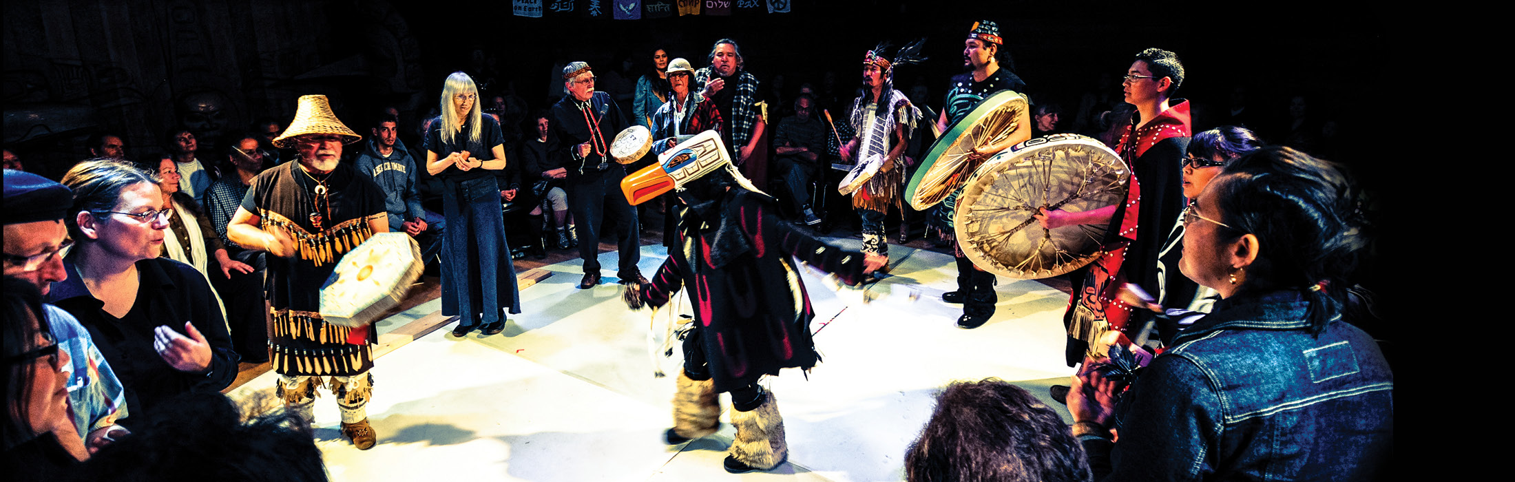 Un public assis regarde une personne se tenir au centre de la scène dans un costume d'oiseau, entourée de personnes vêtues de vêtements traditionnels et tenant des tambours ronds. 