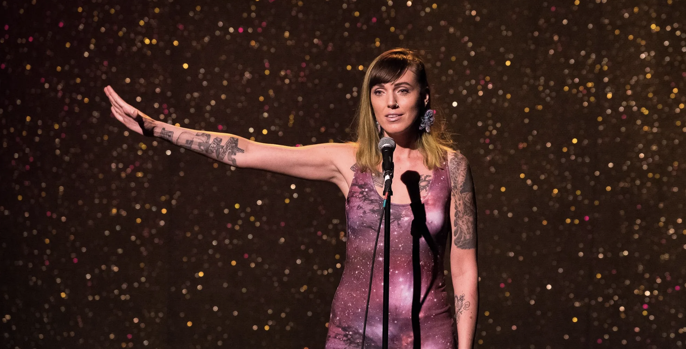 Une personne se tient debout devant un fond chatoyant. Son bras droit est levé à côté de lui et il parle dans un microphone. 