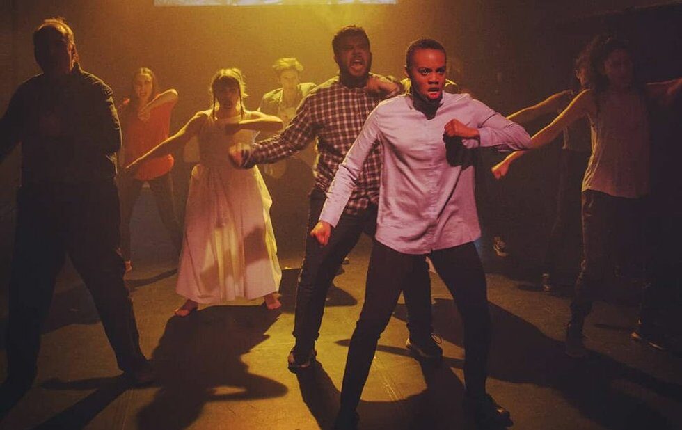 Multiple performers stand on stage holding their left arm up to their chest. Their eyebrows are furrowed and are chanting in unison.