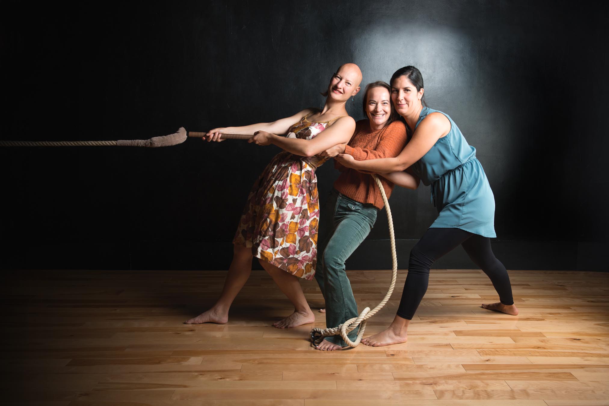 Les trois artistes sont dans une pièce et tirent ensemble une corde épaisse en souriant à la caméra. 