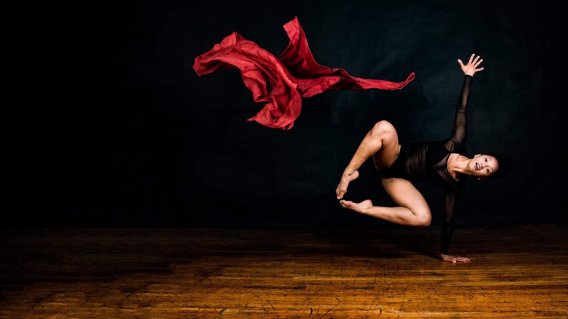 Une personne saute à côté d'eux. Ses jambes et un bras sont en l'air tandis qu'elle se tient en équilibre sur l'autre main. Un grand morceau de tissu rouge en satin flotte dans l'air à côté d'eux. 