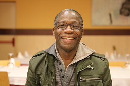 Portrait d'un homme assis dans un réfectoire, souriant, la bouche ouverte, en direction de l'appareil photo. 