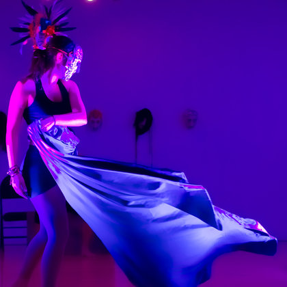 Un personnage masqué se tient dans une pièce à l'éclairage violet et déplace sa cape devant lui.  Derrière lui, trois masques différents sont fixés au mur.