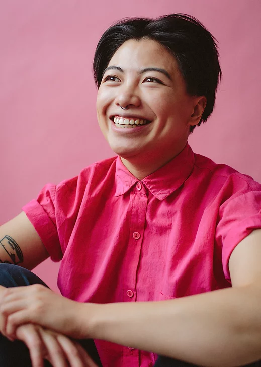Portrait d'une personne assise devant un mur rose et portant une chemise rose. Elle sourit en s'éloignant de l'appareil photo et enroule ses mains autour de ses genoux. 