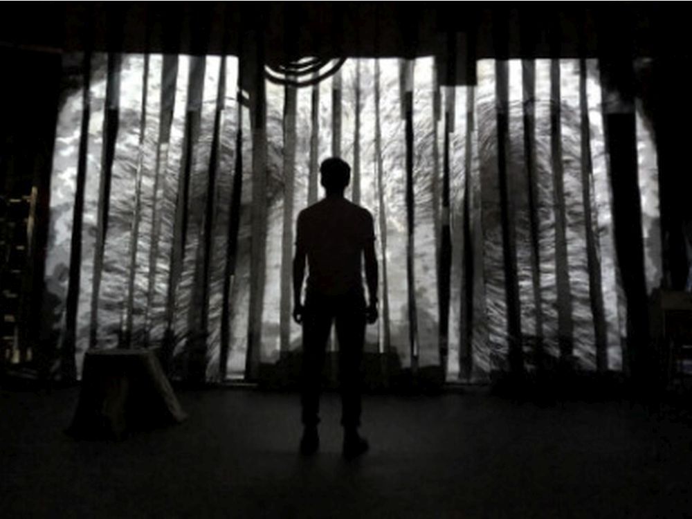 Image en noir et blanc d'un artiste debout au centre d'une scène. Derrière lui se trouve une projection d'une image fragmentée avec des tourbillons de noir et de blanc. Le corps de la personne est rétroéclairé par la projection tandis que l'avant de son corps est recouvert d'une ombre.