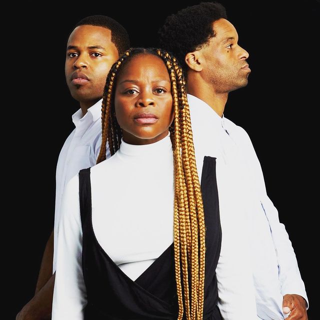 Dans une pièce noire et vide, trois interprètes se tiennent en formation triangulaire, les visages sans expression. La femme à l'avant porte un col roulé blanc à manches longues sous une robe portefeuille noire sans manches et regarde fixement vers la caméra. Les deux hommes sont positionnés derrière ses épaules gauche et droite et portent des chemises à col blanc à manches longues. L'homme de gauche regarde légèrement à gauche de la caméra et l'homme de droite regarde directement devant lui. 